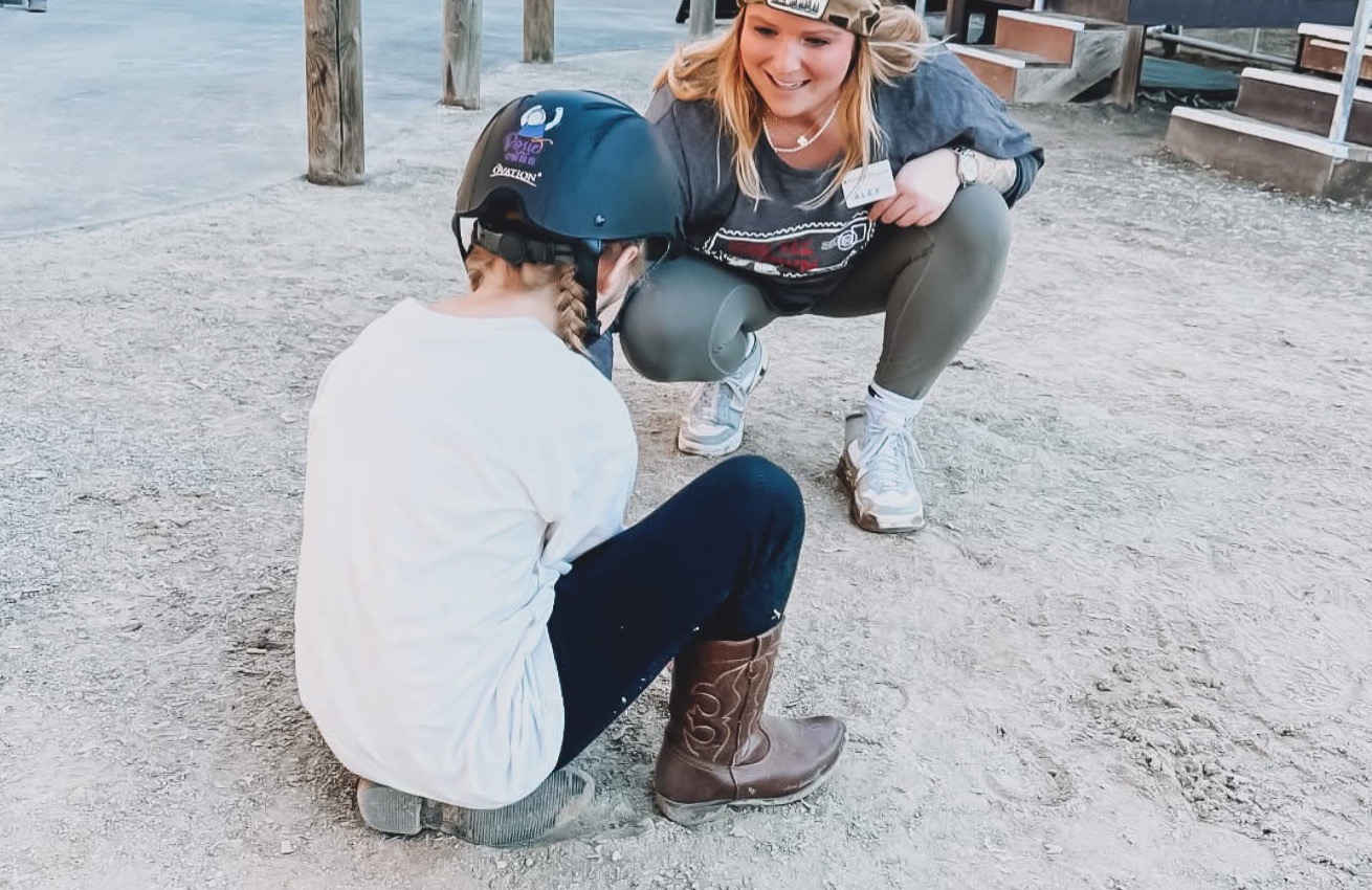 Alexandra kneeling beside a young child wearing a riding helmet during a stable-based session